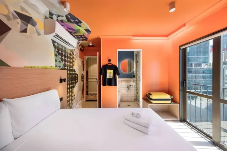 Chambre privée orange avec lit double et grande fenêtre au JO&JOE Auckland