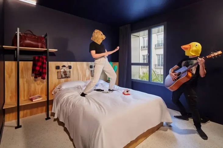 Chambre avec lit double et deux personnes masquées, dont une joue de la guitare, au JO&JOE Paris Nation