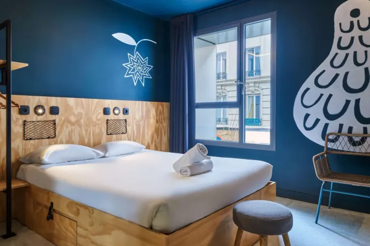 Chambre double avec mur bleu, tête de lit en bois et fenêtre au JO&JOE Paris Nation
